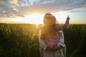 A mother holds her young child in her arms while standing in a lush green field at sunset. The child, dressed in a pink coat and beanie, reaches out toward the golden sky, creating a warm and serene moment.