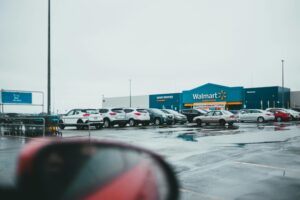 A Walmart Supercentre with a full parking lot on a rainy day. The store's blue and yellow signage is visible in the background, along with shopping carts and parked cars. A car's side mirror is partially visible in the foreground, reflecting part of the scene.