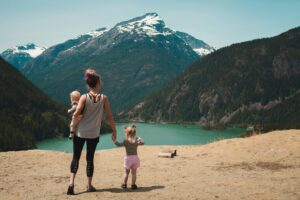 A mother stands on a scenic mountain overlook, holding her baby in one arm and holding hands with her young child. They face a stunning view of a turquoise lake surrounded by lush green mountains with snow-capped peaks in the background.