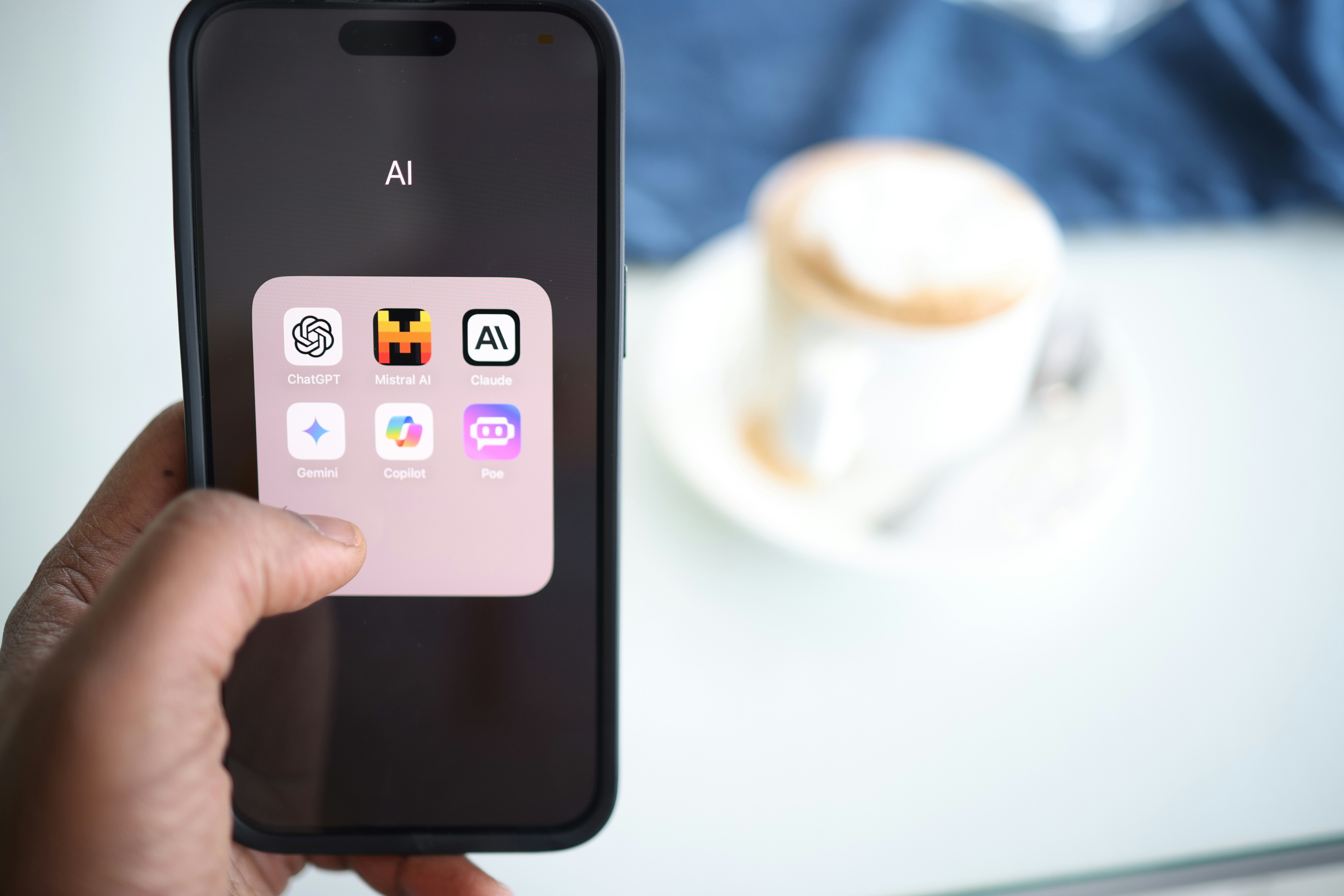 A hand holding a smartphone displaying a folder labeled "AI" with six popular artificial intelligence apps: ChatGPT, Mistral AI, Claude, Gemini, Copilot, and Poe. In the blurred background, there is a cup of cappuccino on a white saucer and a blue cloth.